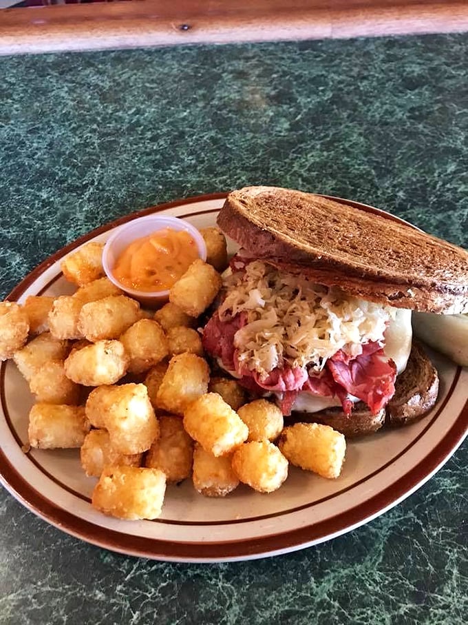 The Reuben: a towering monument to sandwich perfection, flanked by golden tater tots standing at crispy attention.